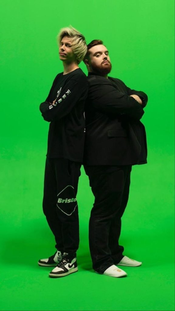 Dos personas de pie, El Rubius e Ibai Llanos, espalda con espalda, posan con actitud desafiante frente a un fondo verde, representando un shipp popular entre streamers