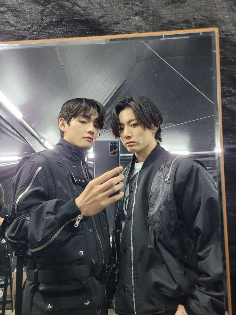 Dos personas, Tehyung y Jungkook de BTS, vestidos de negro se toman una selfie frente a un espejo, reflejando cercanía y estilo coordinado que alimenta el shipp entre ambos.